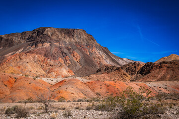 Vivid layered desert mountains in red, orange and tan hues rise under a deep blue sky, sunlit slopes and sparse sagebrush form a dramatic textured landscape.