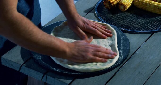 Hand of chef stretching out pizza dough on platform near cooked corn on the cob
