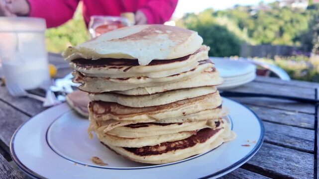 Tall stack of freshly made pancakes sitting on a plate during an outdoor breakfast. A person in the background prepares to eat