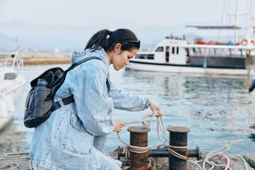 Woman with backpack handling rope at a marina dock, tying lines to bollards for mooring, outdoor...