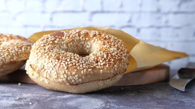 Freshly baked sesame bagels on wooden board with paper wrap and knife in slow motion