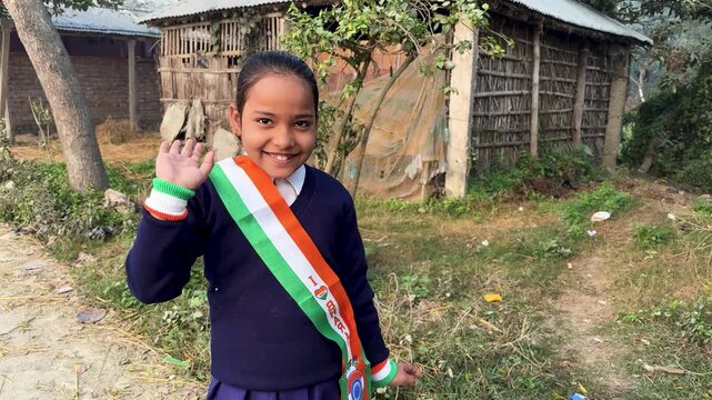 smiling indian school girl waving with tricolor sash in rural village background