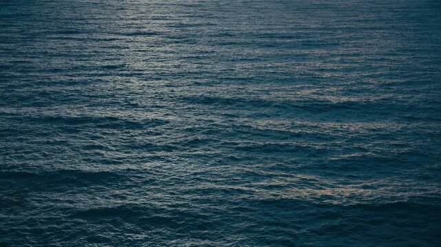 Abstract slow motion view of deep blue ocean waves rolling gently across the surface, subtle ripples and shifting light creating a calm, textured seascape.