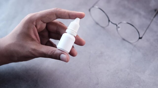 Person holding small white plastic bottle of eye drops over stone surface background near eyeglasses