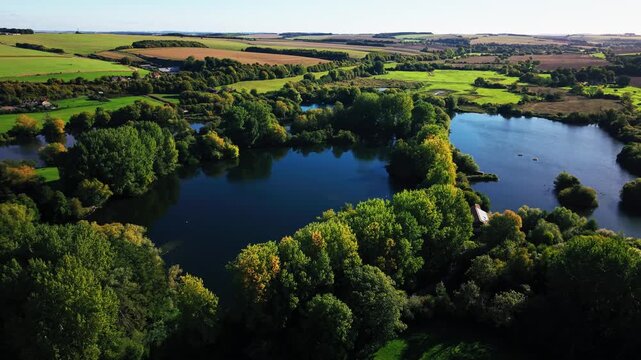 Drone aerial view of lakes surrounded by farmland and woodland in rural Wiltshire, England. Patchwork fields, trees, and calm water create a scenic landscape typical of the British countryside