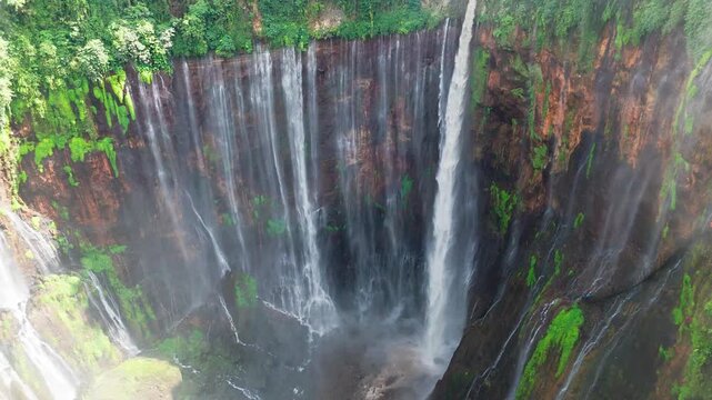 4K drone flying backward above Tumpak Sewu waterfall in East Java, Indonesia, revealing the dramatic semicircle canyon with mist and lush cliffs.