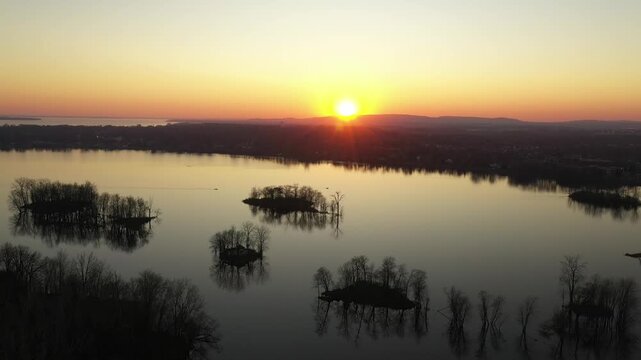 Drone shot, descending gently over calm water, behind the silhouette of a bare tree, during a sunset in a cloudless sky