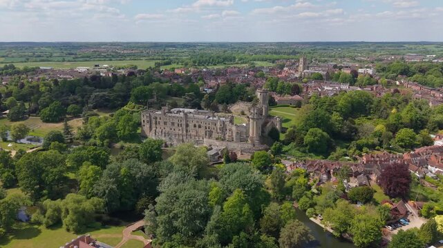 Drone orbit over Warwick village in spring, showcasing lush green landscapes and the historic castle at the center, surrounded by charming countryside and vibrant seasonal scenery.