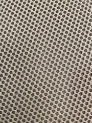Close-up of perforated mesh fabric with repeating square pattern in beige and dark gray tones. The textured textile surface shows small grid openings forming a geometric design, commonly 