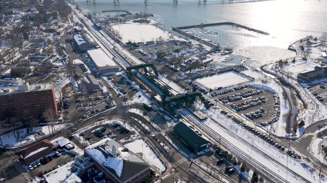 High angle drone view of a snowy commuter train station, platforms and tracks next to a packed parking lot by a frozen river