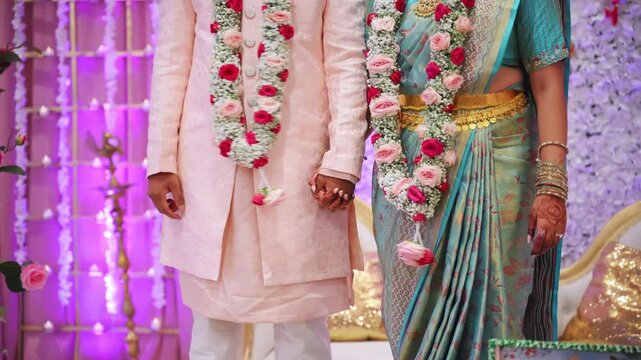 Indian bride and groom holding hands during traditional wedding ceremony. Elegant couple in cultural attire with floral garlands standing together on decorated stage.