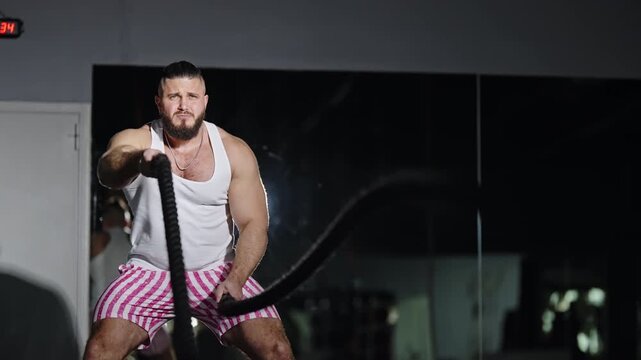 Muscular man powering through intense battle rope workout in an indoor gym, creating powerful rope waves to build strength, endurance, and core stability under dramatic lighting