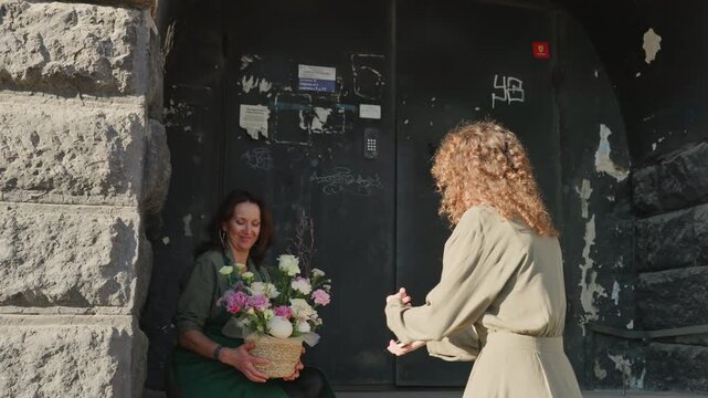 Woman receiving bouquet on street corner as customer hands pastel arrangement to florist seated in stone doorway, warm sunlight illuminates peony and rose bouquet, candid exchange of kindness, basket