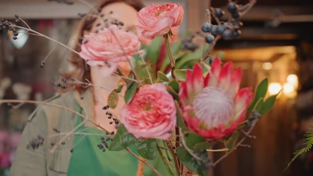 White florist inspecting pink protea bouquet up close, contemplative expression while checking stem health and petal texture in cozy flower shop, warm lights, rustic shelves and ribbon rolls, hands
