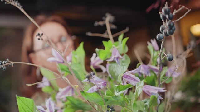 Woman florist arranging purple clematis bouquet in cozy workshop, soft bokeh lights, closeup on stems and leaves, candid portrait with gentle smile, trimming branches into rustic vase, spring floral