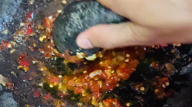Hand grinding fresh chili and spices using traditional stone mortar and pestle to make Indonesian sambal