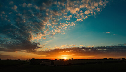 Dramatic sunset paints sky with fiery orange and deep blue hues clouds catch golden light casting long shadows over silhouetted rural landscape vibrant colors creating a breathtaking natural spectacle