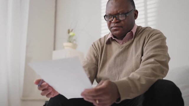Middle-aged black man reading letters from bank or tax service at home interior. Portrait of african american citizen checking utility bills, male person checking agreement, professional lawyer
