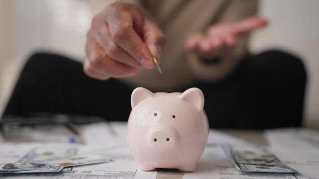 Funny piggy bank on table with utility bills and dollar banknotes, save money. Monthly credit payments and expenses, african american male person putting coins into money-box, financial literacy