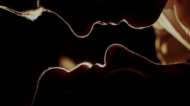 Close silhouette of loving couple sharing tender kiss in warm backlight, intimate relationship and passion concept, emotional connection in dark dramatic lighting.