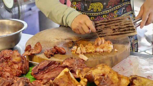 Close-up footage of a Thai street food vendor slicing grilled pork on a wooden chopping board using a large Chinese cleaver. Authentic market atmosphere and traditional food preparation in Thailand.