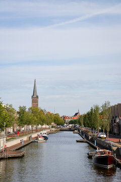 Canal with boats and residential houses along riverside street, calm urban scene. River with church tower in background. Vertical portrait photo.