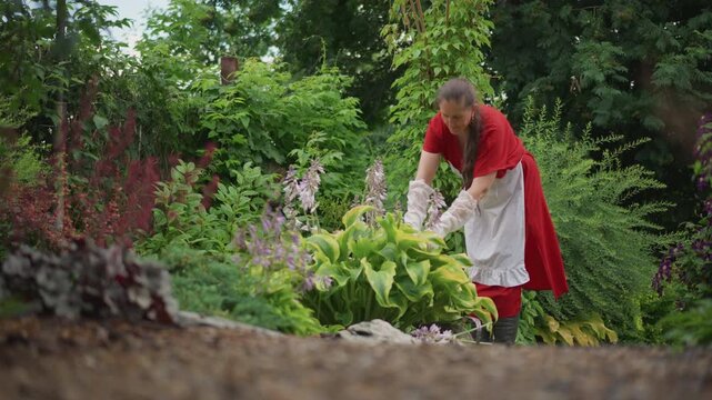 White woman caretaker gathering plants near hosta border, arranging leaves and blooms into gentle display, warm expressive smile, steady hands in red dress and apron, uplifting nurturing scene in lush