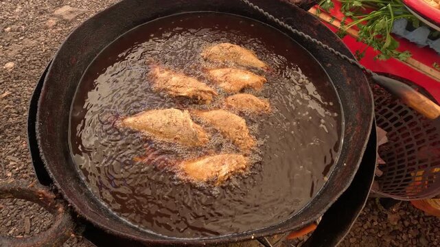 Breaded fish fillets frying in hot oil, turning golden brown with bubbling crust. Classic street-style fish preparation outdoors, showing crispy coating forming in an active frying setup