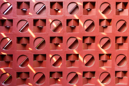 Seamless geometric pattern of a red terracotta screen wall. Sunlight casts beautiful shadows on the hollow ventilation blocks, creating a warm abstract architectural texture.