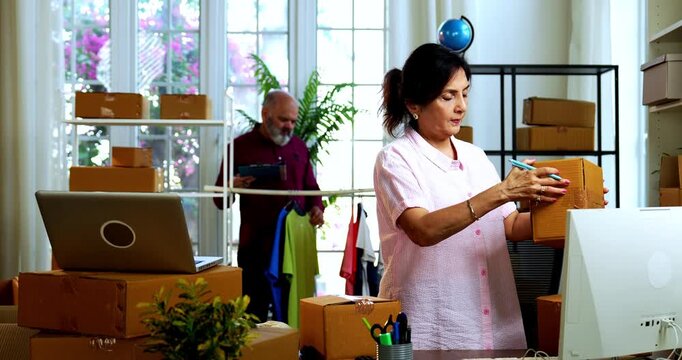 Indian senior couple doing E-commerce business from home, busy working in office managing online orders, packing parcels, dispatching boxes, taking customer calls and handling inventory together