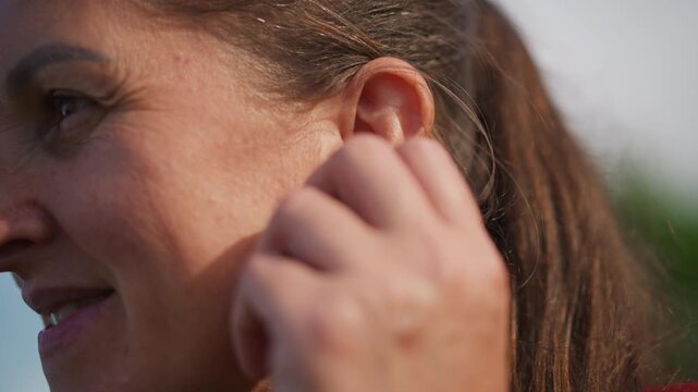 Closeup of lady adjusting earring during sunset. Gentle portrait of woman with ponytail in warm sunlight. Calm outdoor image capturing woman softly smiling amidst golden sunset light