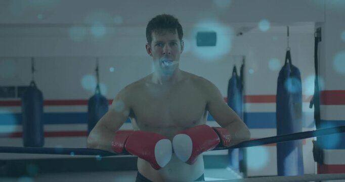 Male boxer leaning on ring ropes, breathing through mouthguard and resting, blue bokeh obscuring