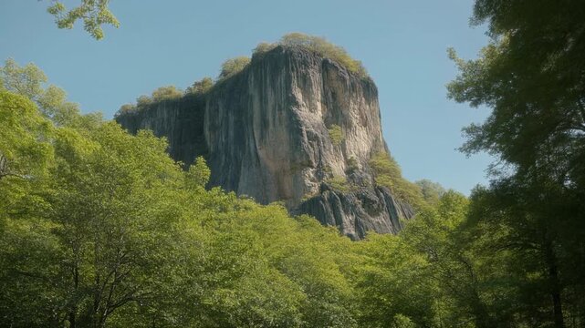 A tall rock formation with a flat top emerges behind a vibrant canopy of green trees beneath a cloudless blue sky
