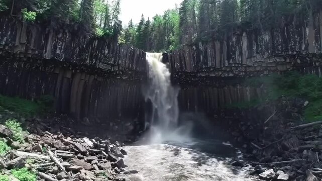 A tall waterfall cascades down a columnar basalt cliff, surrounded by lush green trees. Water rushes over rocks below, forming rapids