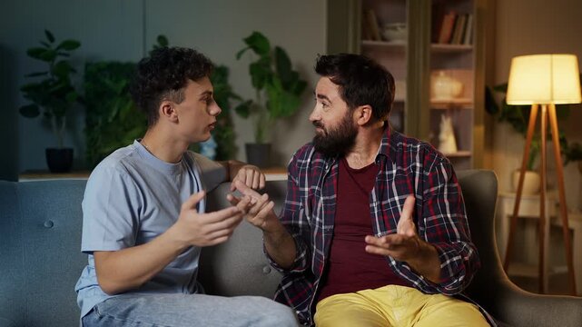 Happy father communicating with adolescent son at home, telling funny stories