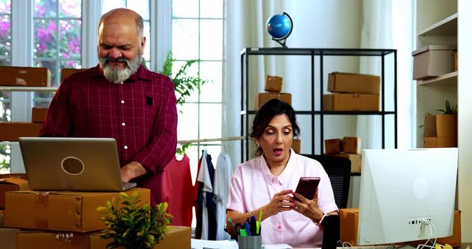 Indian senior couple celebrating success in home based E-commerce business while managing online orders, dispatching parcels, packing cardboard boxes and working together in home office