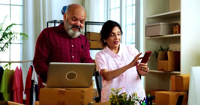 Indian senior couple celebrating success in home based E-commerce business while managing online orders, dispatching parcels, packing cardboard boxes and working together in home office