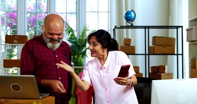 Indian senior couple celebrating success in home based E-commerce business while managing online orders, dispatching parcels, packing cardboard boxes and working together in home office