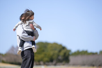 A man is holding a child in his arms. The child is wearing a gray shirt and black pants. The scene is outdoors, with a blue sky in the background. Scene is warm and loving