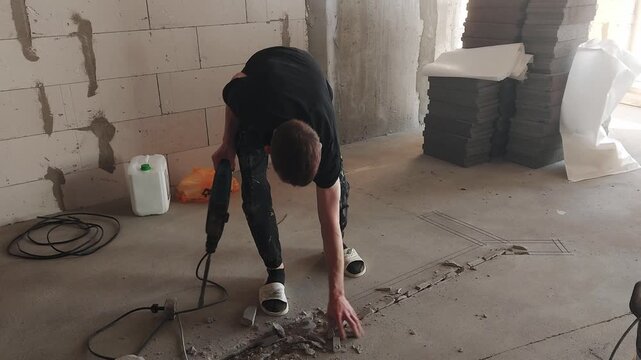 A worker uses a hammer drill to break up a concrete floor at a construction site. Construction materials and tools are scattered around. It's daytime, and the worker is focused.