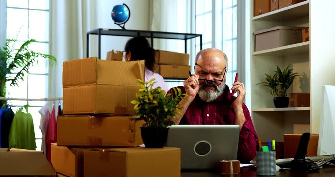 Indian senior couple enjoying working, taking online orders, phone call while doing E-commerce business from home managing parcels, packing boxes and handling customer communication happily