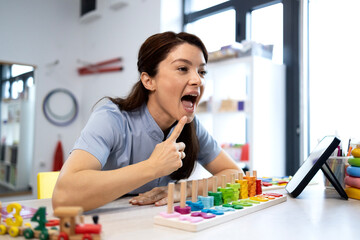 Fototapeta premium Speech therapist demonstrating oral motor exercises while providing teletherapy services, using a tablet and educational toys for children's speech development and articulation training