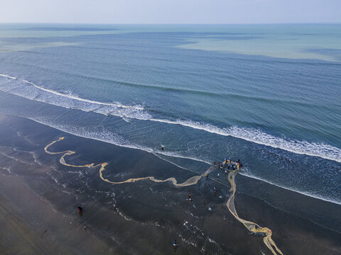 Cox's Bazar, Bangladesh - 16 May 2024: Aerial view of the dark sandy beach contrasting sharply with the turquoise sea, as fishermen haul in their nets, creating winding patterns on the shore.