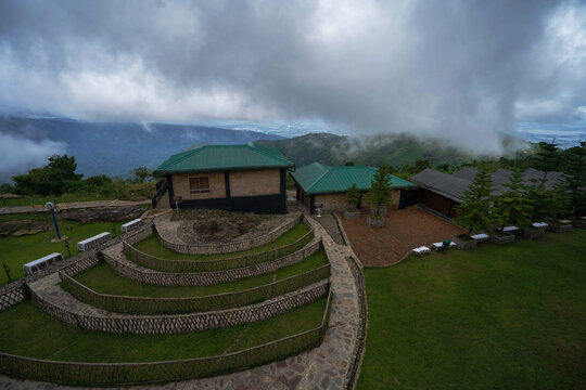 Cherrapunji, India - 26 July 2023: View of a serene landscape where the green roofs of buildings nestle amidst rolling hills cloaked in mist, under a sky heavy with clouds.