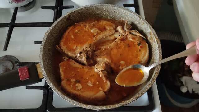 Person basting three pork pieces simmering in red sauce on frying pan with spoon, first person view video