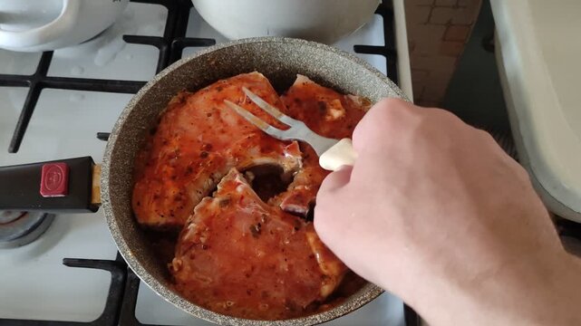 Person pressing and testing three pork pieces simmering in red sauce on skillet with double fork to check doneness, first person view video with sound
