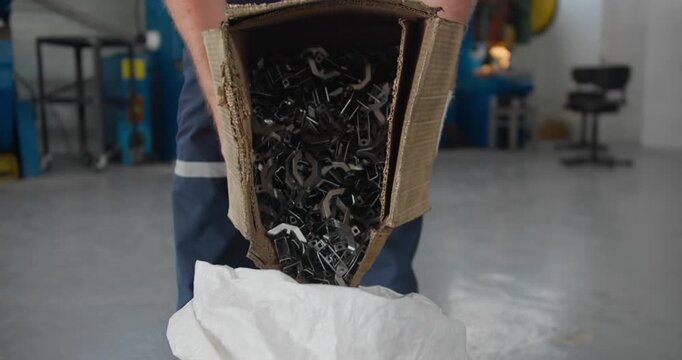 Worker pouring stamped metal parts from cardboard box into container at factory floor. Industrial manufacturing workflow, post-press handling
