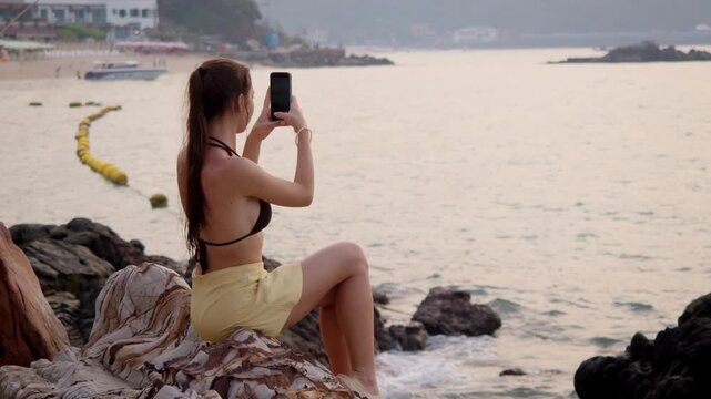 Attractive young woman with toned figure photographs sunset seascape from boulder perch holding smartphone vertical. Yellow skirt contrasts rocky terrain while buoys dot calm water ahead.