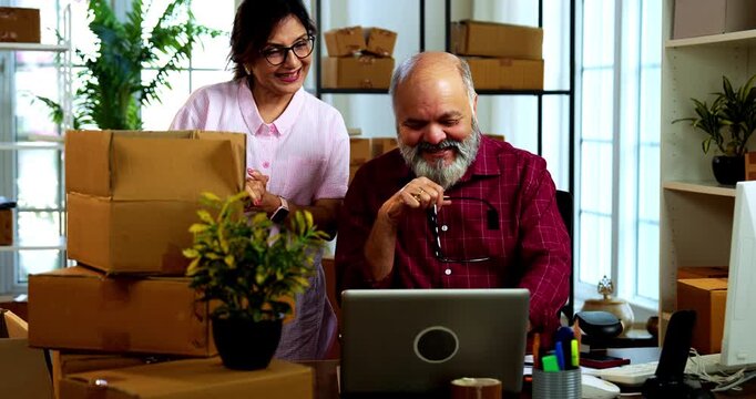 Indian senior couple celebrating success in home based E-commerce business while managing online orders, dispatching parcels, packing cardboard boxes and working together in home office