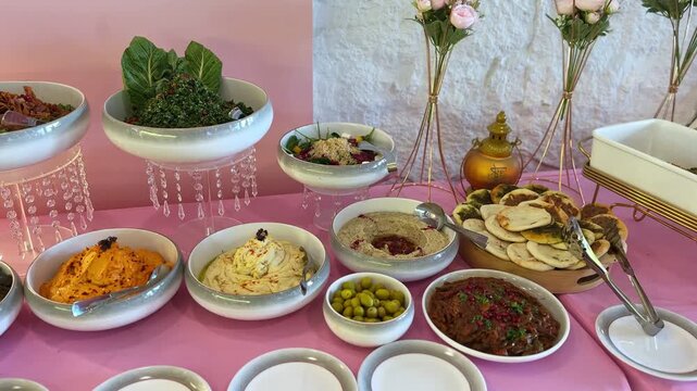 Wide view of a Middle Eastern mezze buffet featuring hummus, creamy dips, tabbouleh salad, olives and stacked flatbread arranged on a decorated indoor catering table with elegant presentation.